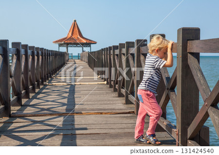 People walk by long wooden pier with traditional gazebos stretches over calm turquoise sea under clear blue sky. Perfect tropical scenery for travel, resort, summer vacation, coastal tourism themes. 131424540