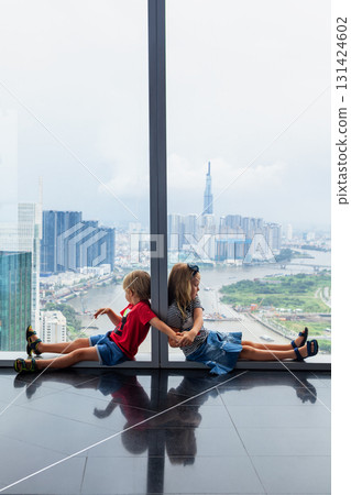 Funny kids look at panoramic view of Ho Chi Minh City from modern skyscrapers observation deck. Urban lifestyle, travel with kids, and high-rise cityscape concept with Southeast Asian charm. 131424602