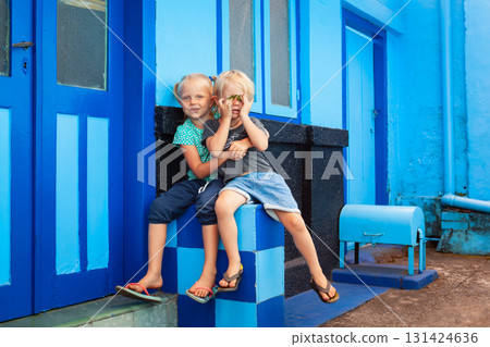 Happy kids sitting outside a vibrant blue-painted house in Kampung Biru Arema, Malang, Java, Indonesia. Colorful urban setting perfect for promoting family travel lifestyle, Southeast Asian culture. Happy kids sitting outside a vibrant blue-painted house in Kampung Biru Arema, Malang, Java, Indonesia. Colorful urban setting perfect for promoting family travel lifestyle, Southeast Asian culture. 131424636