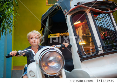 Smiling boy sits on a retro scooter museum. Funny moment during a family trip. Perfect for themes of travel on vacation, adventure, exploring, vintage vehicles, museums, family tourism. 131424639