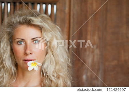 Young blonde woman with frangipani flower on open-air veranda of wooden bungalow in tropical garden. Girl relaxing in luxury villa on family summer vacation. Beach travel, spa, natural lifestyle. Young blonde woman with frangipani flower on open-air veranda of wooden bungalow in tropical garden. Girl relaxing in luxury villa on family summer vacation. Beach travel, spa, natural lifestyle. 131424697