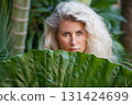 Blonde woman hiding behind a giant tropical leaf in a lush jungle in Bali. Natural portrait that evokes travel, wellness, connection with nature. Ideal for spa, retreats, summer travel lifestyle. 131424699