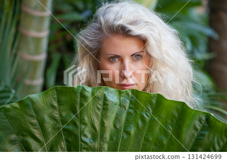 Blonde woman hiding behind a giant tropical leaf in a lush jungle in Bali. Natural portrait that evokes travel, wellness, connection with nature. Ideal for spa, retreats, summer travel lifestyle. Blonde woman hiding behind a giant tropical leaf in a lush jungle in Bali. Natural portrait that evokes travel, wellness, connection with nature. Ideal for spa, retreats, summer travel lifestyle. 131424699