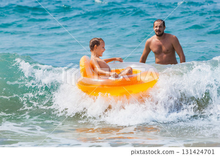 Happy kid with father have fun in sea surf on beach. Joyful child on inflatable ring ride on breaking wave. Travel lifestyle, swimming activities in family summer camp. Vacations on tropical island Happy kid with father have fun in sea surf on beach. Joyful child on inflatable ring ride on breaking wave. Travel lifestyle, swimming activities in family summer camp. Vacations on tropical island 131424701