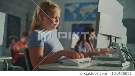 Blonde Girl in Blue Shirt Works on Desktop Computer While Classmates Sit at Desks 131426091