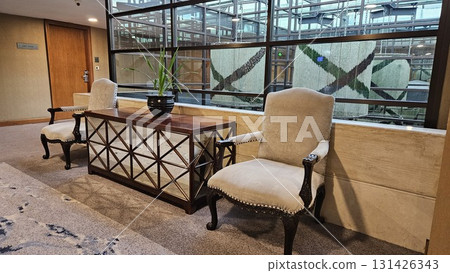 A pair of elegant upholstered armchairs and a mirrored console table with a potted plant create a sophisticated seating area in a modern hallway with large glass windows. A pair of elegant upholstered armchairs and a mirrored console table with a potted plant create a sophisticated seating area in a modern hallway with large glass windows. 131426343
