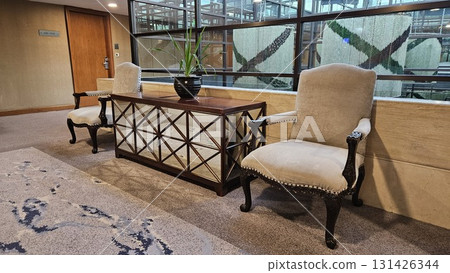 A pair of elegant upholstered armchairs and a mirrored console table with a potted plant create a sophisticated seating area in a modern hallway with large glass windows. 131426344