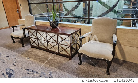 A pair of elegant upholstered armchairs and a mirrored console table with a potted plant create a sophisticated seating area in a modern hallway with large glass windows. A pair of elegant upholstered armchairs and a mirrored console table with a potted plant create a sophisticated seating area in a modern hallway with large glass windows. 131426345