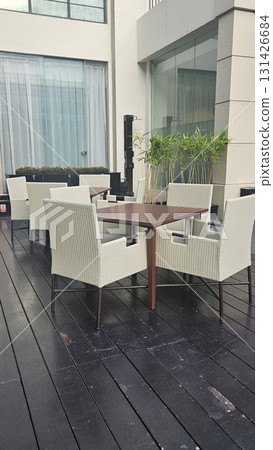 A tranquil outdoor dining area features several modern tables and wicker chairs arranged on a dark wooden deck, surrounded by lush green plants and a contemporary building. A tranquil outdoor dining area features several modern tables and wicker chairs arranged on a dark wooden deck, surrounded by lush green plants and a contemporary building. 131426684