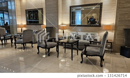 A luxurious hotel lobby features a classic, elegant seating area with plush gray armchairs, a comfortable sofa, and a glass coffee table, all set against a backdrop of marble walls and large ornate mi 131426714