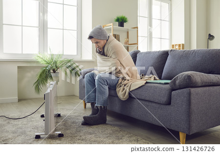 Man sitting on sofa at home wrapped in blanket and trying to warm up on electric heater. 131426726
