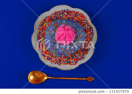On the plate is a plate with a blue background and a pink glazed donut with sprinkles on top. A gold spoon is next to the plate On the plate is a plate with a blue background and a pink glazed donut with sprinkles on top. A gold spoon is next to the plate 131427979
