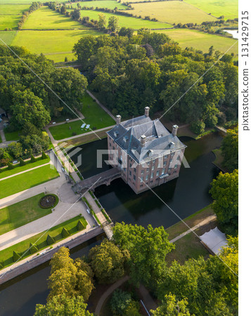 Aerial view of historic moated castle with gardens, bridge, and adjacent long building, surrounded by trees, water features, and a nearby village under warm sunlight. 131429715