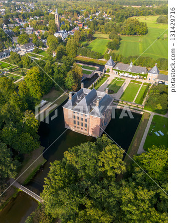 Aerial view of historic moated castle with gardens, bridge, and adjacent long building, surrounded by trees, water features, and a nearby village under warm sunlight. 131429716