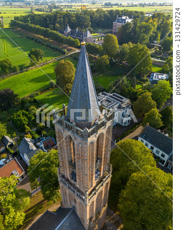 Aerial view of Gothic brick church tower with pointed roof and pinnacles, surrounded by trees and residential houses in a quiet neighborhood. 131429724