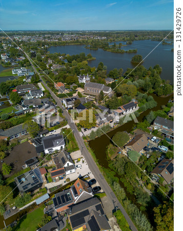 Aerial view of a small town with a central church and tall steeple, surrounded by houses, tree lined streets, and nearby lakes with islands, blending historic charm and natural beauty. 131429726