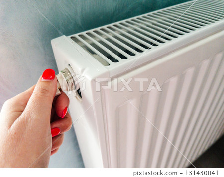 Close-up of a womans hand adjusting a white panel radiator valve at home. Concept of winter heating, temperature control, and energy efficiency. Maintenance action to bleed air, boost heat output 131430041
