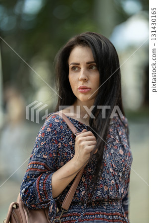 woman with long dark hair wears a floral patterned dress while standing in a park. She holds a bag and gazes off into the distance enjoying a warm day. 131430165