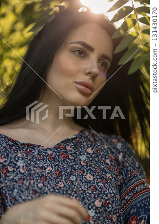 young woman stands gracefully among lush green leaves wearing a floral dress. Sunlight shines through the foliage highlighting her features as she gazes thoughtfully into the distance. 131430170