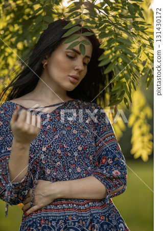 woman with dark hair stands thoughtfully among lush green leaves. She wears a colorful dress and enjoys the peaceful atmosphere created by the warm sunlight filtering through the trees. 131430172