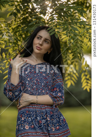 In a peaceful park a young woman stands elegantly beneath a tree with vibrant green leaves. 131430173