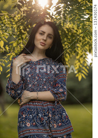 woman stands confidently in a colorful floral dress captured during the golden hour in a lush garden. Soft sunlight filters through the trees creating a serene atmosphere. 131430174
