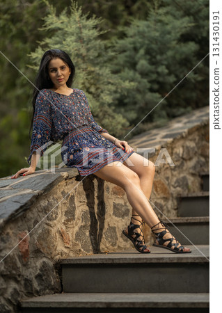 woman with long hair sits casually on stone steps wearing a colorful patterned dress and stylish sandals. Lush greenery surrounds her creating a peaceful atmosphere in the evening. 131430191