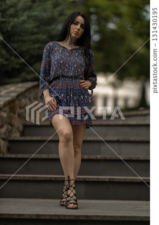 young woman confidently walks down a set of stone steps. She wears a colorful dress and sandals surrounded by greenery in a peaceful garden setting. 131430195