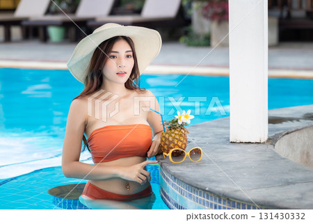 Sexy young asian woman in swimsuits in summer. beautiful woman in orange bikini with hat and tropical drink in swimming pool. 131430332