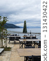 An outdoor dining area with wooden tables and black chairs arranged in rows, overlooking a serene sea with mountains in the background. 131430422