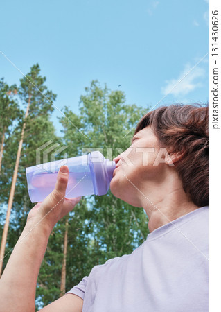 Mature woman drinks water from bottle after workout against background of forest and sky 131430626