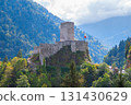 An ancient castle with a stone tower and walls, surrounded by lush greenery and trees, is situated on a rocky hill in a mountainous area. The castle is built with rough stone and has a red flag waving 131430629