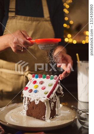 Person Decorating Gingerbread House With Powdered Sugar In Festive Home Kitchen Person Decorating Gingerbread House With Powdered Sugar In Festive Home Kitchen 131430960