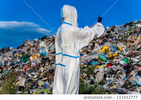 Individual wearing a protective suit captures a moment at a large garbage dump, surrounded by heaps of waste under a clear sky Individual wearing a protective suit captures a moment at a large garbage dump, surrounded by heaps of waste under a clear sky 131432777