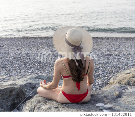 Attractive woman with long hair, wearing red bikini and straw hat, on the beach, sitting on a large rock, sunbathing. 131432911