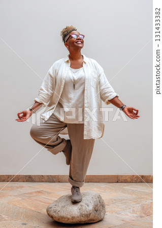 Joyful plus-size Black woman practicing yoga indoors standing on stone in tree pose 131433382