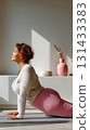 Mature plus-size woman doing yoga in cobra pose on mat indoors, surrounded by natural light and decorative elements. 131433383