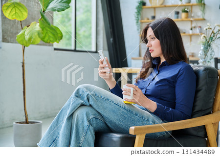 Serious sad young woman sitting at home in chair using smartphone Serious sad young woman sitting at home in chair using smartphone 131434489