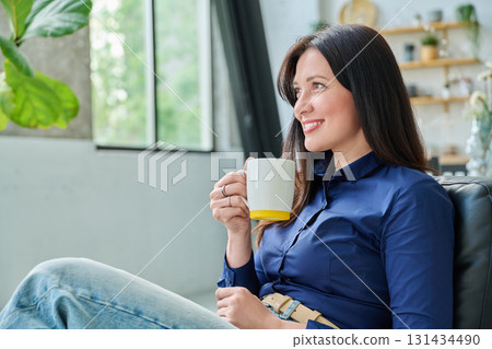 Portrait of beautiful 30s smiling woman with cup sitting in armchair at home in living room Portrait of beautiful 30s smiling woman with cup sitting in armchair at home in living room 131434490