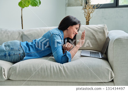 Young woman looking at laptop screen talking having video call lying on couch at home Young woman looking at laptop screen talking having video call lying on couch at home 131434497