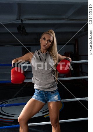blonde woman dons red boxing gloves and a gray athletic top while posing confidently in a boxing ring. She seems prepared for training showcasing strength and focus. 131435119