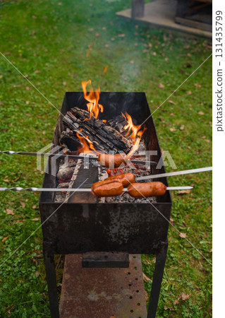 Grilling sausages over open fire on metal barbecue grill in backyard with green grass in springtime Grilling sausages over open fire on metal barbecue grill in backyard with green grass in springtime 131435799