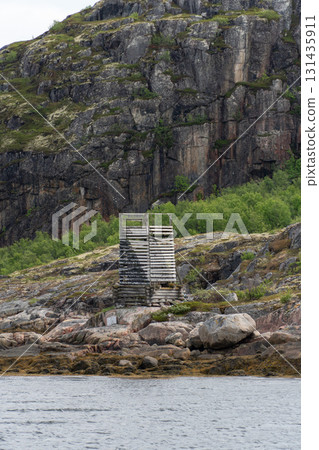 Old wooden coastal structure stands on rocky shore against steep cliffs with moss and greenery 131435911