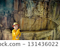 A young boy in a yellow hat and shirt is leisurely pointing at an ancient statue made of wood. This sculpture depicts an important visual arts piece in rock 131436022