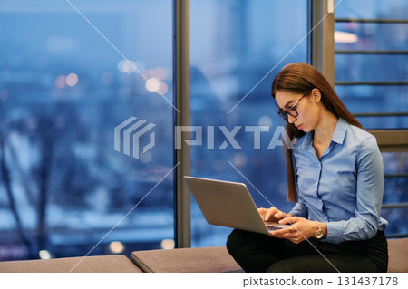 A businesswoman utilizes her laptop while seated by the window of a large corporate building, offering a picturesque view of the city skyline as her backdrop. A businesswoman utilizes her laptop while seated by the window of a large corporate building, offering a picturesque view of the city skyline as her backdrop. 131437178