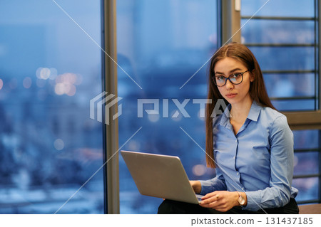 A businesswoman utilizes her laptop while seated by the window of a large corporate building, offering a picturesque view of the city skyline as her backdrop. 131437185