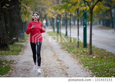 Young beautiful woman running in autumn park and listening to music with headphones on smartphone 131437233