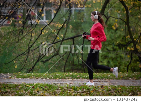 Young beautiful woman running in autumn park and listening to music with headphones on smartphone 131437242