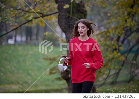 Young beautiful woman running in autumn park and listening to music with headphones on smartphone Young beautiful woman running in autumn park and listening to music with headphones on smartphone 131437265