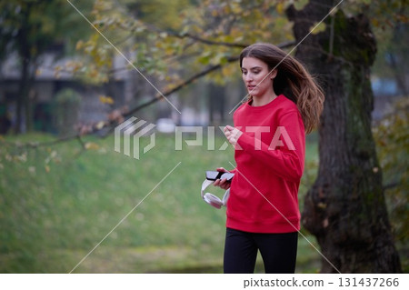 Young beautiful woman running in autumn park and listening to music with headphones on smartphone Young beautiful woman running in autumn park and listening to music with headphones on smartphone 131437266